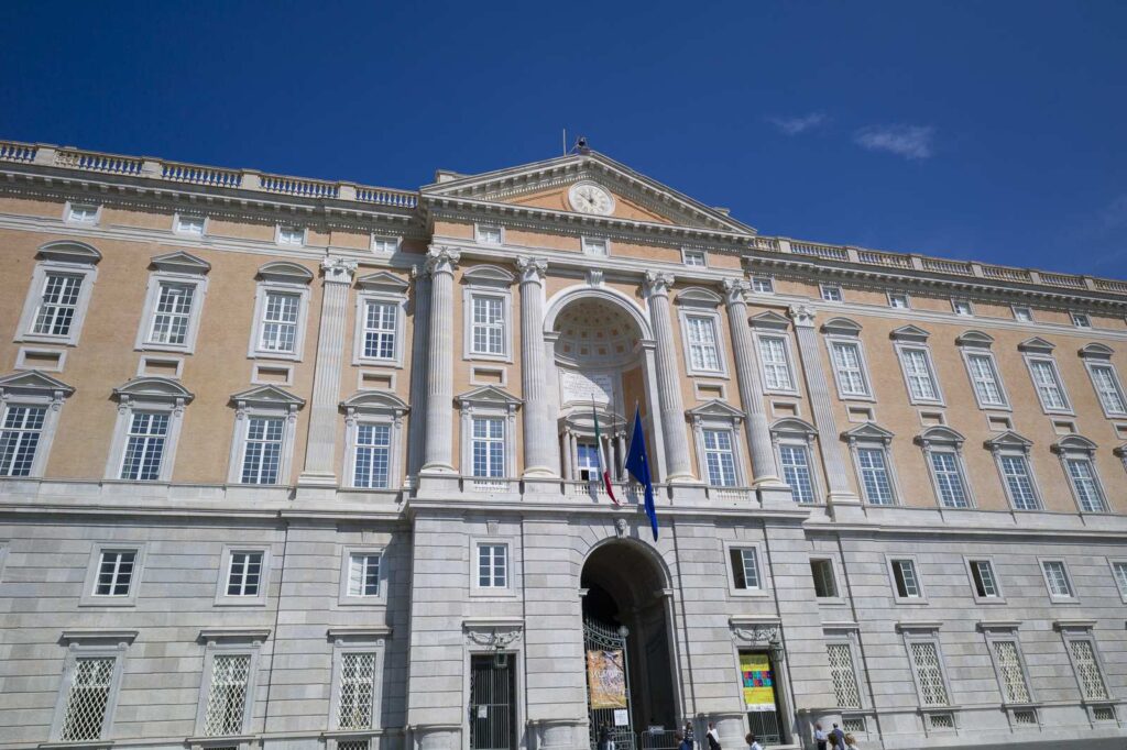 Façade principale du palais royal de Caserte avec colonnes, drapeaux et ciel dégagé.