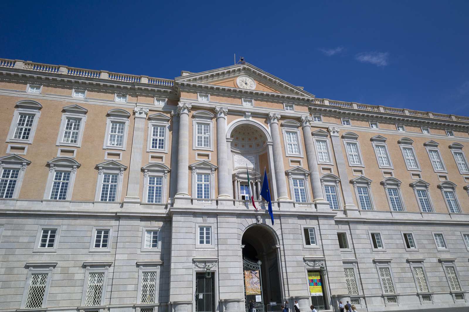 Façade principale du palais royal de Caserte avec colonnes, drapeaux et ciel dégagé.
