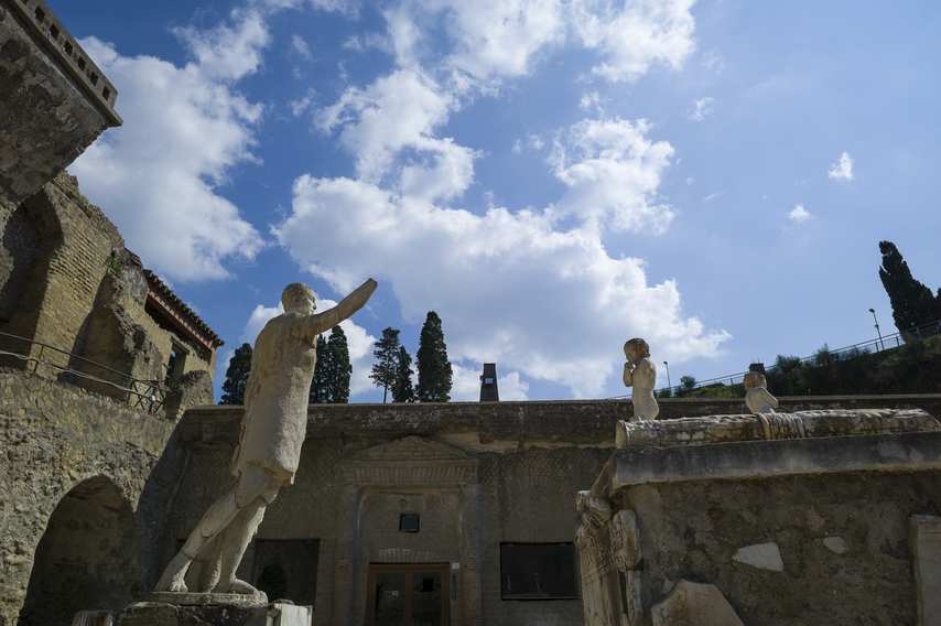 Roman statues at Herculaneum Archaeological Park, one with arm raised, under a blue sky with white clouds